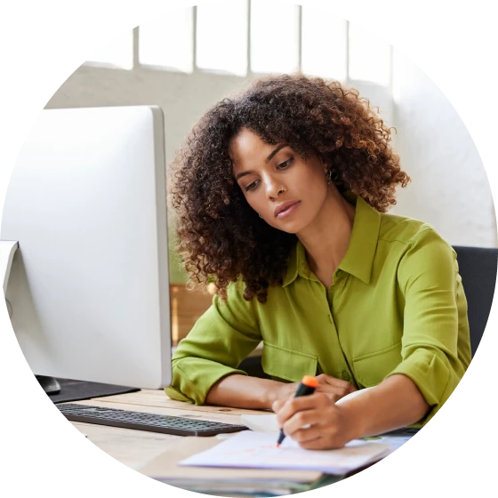 Woman seated behind computer monitor, with pen in hand, concentrating on paperwork